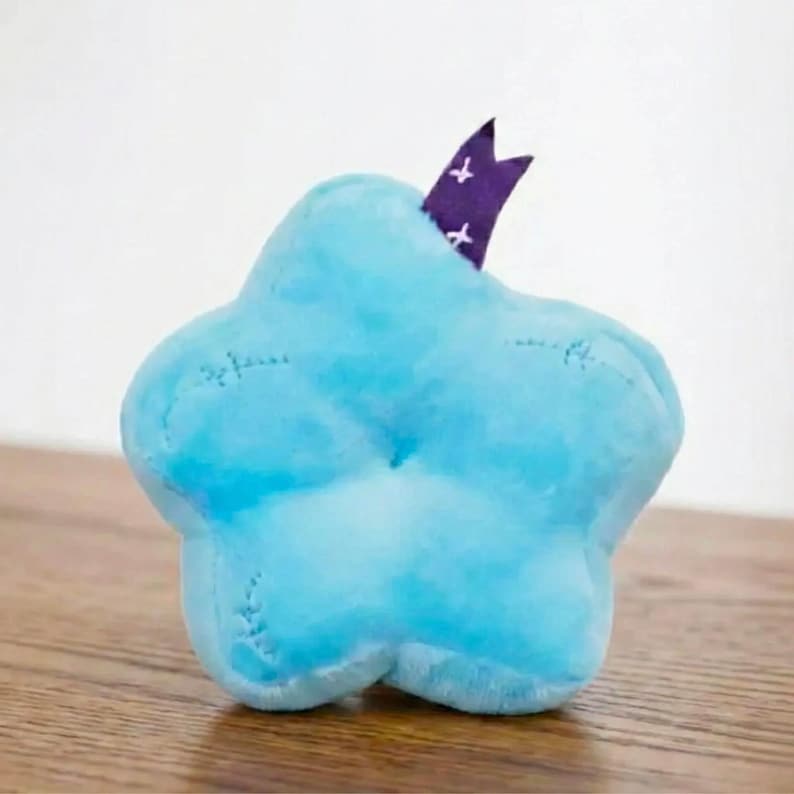 Pu&ograve; includere: Un giocattolo a forma di stella blu chiaro in peluche con dettagli cuciti. Un nastro viola con accenti a croce bianchi &egrave; inserito nella parte superiore. Il giocattolo &egrave; su una superficie di legno.
