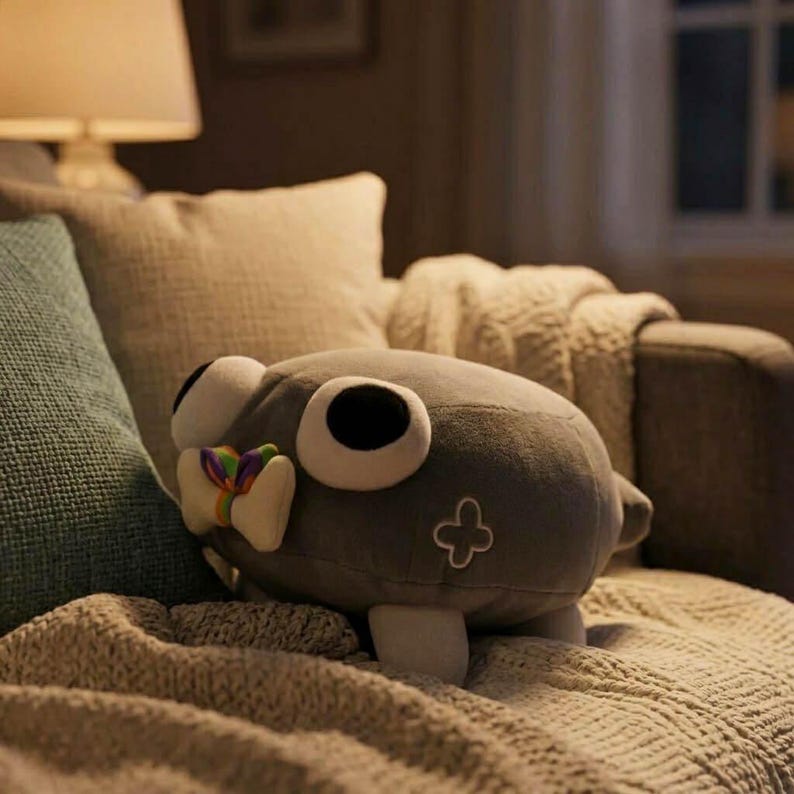 May include: A gray plush toy with large, round black eyes and a white bow with rainbow accents. The stuffed animal has a cross-shaped design on its side and is resting on a textured blanket.