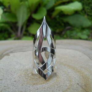 Könnte beinhalten: Ein silberfarbener Ring mit einem aufwendigen, durchbrochenen Design. Der Ring hat eine spitze Spitze und verschlungene Muster, die ein einzigartiges und stilvolles Accessoire schaffen. Der Ring wird auf einer neutralen Oberfläche mit verschwommenem grünem Hintergrund präsentiert.