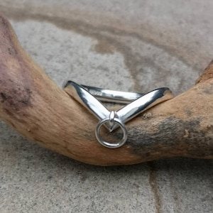 May include: A silver ring with a distinctive V-shaped design and a small circular detail. The ring is presented on a piece of driftwood, emphasizing its minimalist and contemporary style. The ring's polished surface reflects light, highlighting its sleek lines.