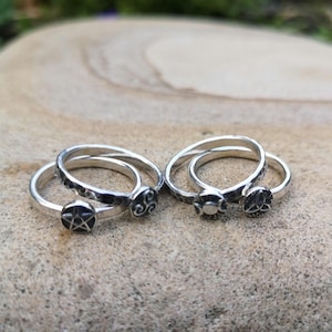 May include: Several silver rings are displayed, each with a unique design. One ring has a pentagram, another a Celtic knot, and others have floral or geometric patterns. The rings are presented on a textured, light brown surface.