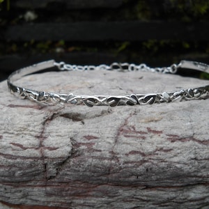 Op de afbeelding: Een zilverkleurige armband met een delicaat, herhalend bladerenontwerp. De armband is dun en flexibel, met een ketting sluiting. Het wordt getoond op een getextureerde, grijze steen, met een vage groene achtergrond.