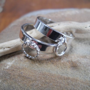 Può includere: Due anelli d'argento, uno con un anello intrecciato e l'altro con un anello semplice, sono impilati. Gli anelli hanno una superficie lucida e riflettente, su uno sfondo neutro.