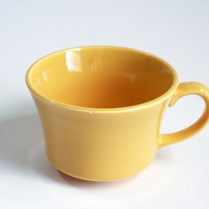 May include: A yellow ceramic teacup with a handle. The cup is empty and sitting on a white surface.
