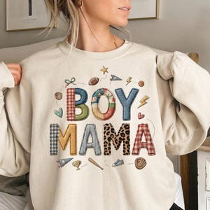 May include: Cream-colored sweatshirt with the words "BOY MAMA" in a mix of patterned and textured fonts. The design includes sports-themed graphics like a football, baseball, and lightning bolt. A cute and cozy top for a mom.