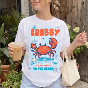 以下が含まれることがあります： オレンジ色のカニのグラフィックと「if Crabby please return to the beach」のテキストが入った白いTシャツ。デザインにはヒトデと貝殻が含まれており、青と白のストライプの背景が特徴です。