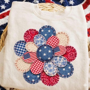 May include: A white sweatshirt featuring a large, appliqued flower design. The flower petals are made from various fabrics in red, white, and blue patterns, including stars, stripes, and floral prints. The design evokes a patriotic theme.