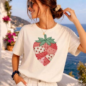 Puede incluir: Camiseta color crema con un diseño de fresa. La fresa está compuesta por varios parches de tela en rojo, rosa y blanco, con un lazo verde a cuadros. El diseño tiene una estética de patchwork.