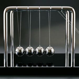 May include: A Newton's cradle, a physics demonstration device, features five polished steel balls suspended in a rectangular frame. One ball is raised and released, transferring momentum to the others. The base is black.