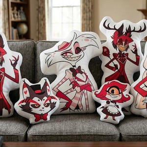 May include: A collection of character-shaped pillows featuring designs from the animated series. The pillows showcase various characters in vibrant red, black, and white colors, arranged on a gray sofa. The pillows are soft and plush.