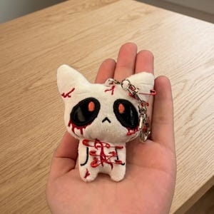 May include: A white plush cat keychain with black eyes, red blood-like accents, and red safety pins. The keychain has a silver chain and clasp. The cat's design has a spooky, Halloween theme.