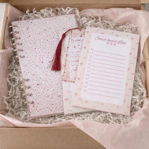 May include: A gift box containing a spiral-bound notebook, a notepad, and a bookmark with a red tassel. The notebook and notepad have a white background with red speckles and the notepad has the text "Sweet heart List". The items are nestled in pink tissue paper and shredded paper.