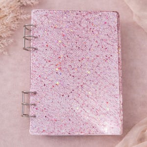 May include: A pink glitter-covered notebook with silver metal binder rings. The notebook has a rectangular shape and is covered in a sparkling glitter pattern. The background is a soft pink color.