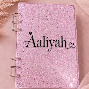 May include: A pink glitter notebook with silver metal rings. The notebook cover features the name "Aaliyah" in black script with a heart above the name and a heart at the end. The notebook is on a pink background.