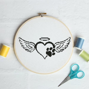 May include: A cross-stitch embroidery hoop with a black heart, paw print, angel wings, and a halo. The design is on white fabric, within a wooden hoop. Yellow, blue, and green thread spools and blue scissors are also visible.