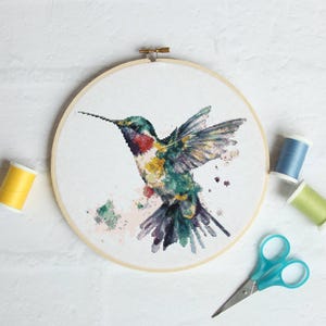 Op de afbeelding: Een kruissteekborduurwerk van een kolibrie in vlucht, met tinten groen, geel, rood en paars. De borduursel is geplaatst in een houten borduurring. Extra items zijn gele, blauwe en groene garenklosjes en een blauwe schaar.