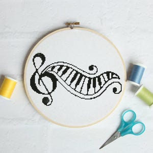 Puede incluir: Diseño de punto de cruz en blanco y negro de una clave de sol y teclas de piano, en un bastidor de bordado de madera. El diseño está rodeado de carretes de hilo amarillo, azul y verde, y unas tijeras azules.