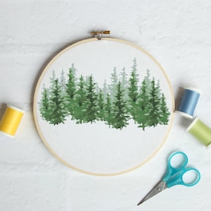 Pode incluir: Um bordado em ponto cruz de uma cena de floresta num bastidor de madeira. O desenho apresenta árvores verdes sobre um fundo branco. Uma bobina de linha amarela, duas outras bobinas de linha e tesouras azuis também estão na imagem.