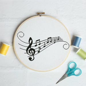 Puede incluir: Un bastidor de bordado de punto de cruz con una clave de sol negra y notas musicales sobre tela blanca. El bastidor de madera está rodeado de carretes de hilo amarillo, azul y verde, y unas tijeras azules.