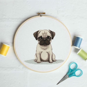 May include: A cross-stitch embroidery of a pug dog in a wooden hoop. The pug is beige and brown, with large dark eyes. Yellow, blue, and green thread spools and blue scissors are arranged around the hoop on a white surface.