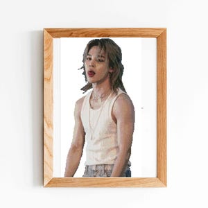 Puede incluir: Un marco de madera rectangular que contiene el retrato de una persona con cabello hasta los hombros, vistiendo una camiseta sin mangas blanca acanalada y un collar fino. La persona mira al frente con la lengua ligeramente fuera, fondo blanco.
