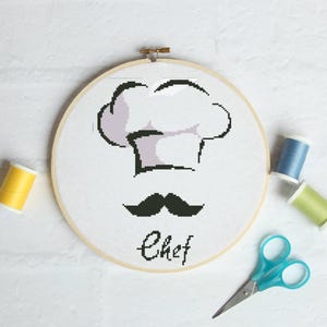 May include: A circular wooden embroidery hoop containing white fabric with a cross-stitch design of a chef hat, a black mustache, and the word Chef in black script. The hoop is surrounded by a yellow spool, a blue spool, a green spool, and scissors.