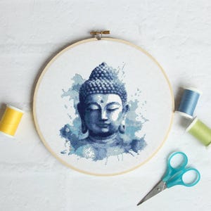 Peut inclure: Broderie au point de croix d'une tête de Bouddha bleue sur un cercle de tissu blanc. Le motif présente un visage et une coiffe détaillés sur un fond aquarelle. Des bobines de fil jaune, bleu et vert et des ciseaux turquoise sont placés autour du cercle.