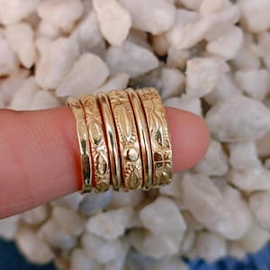 Può includere: Una collezione di anelli color oro con vari disegni in rilievo, tra cui motivi floreali e geometrici. Gli anelli sono impilati, mostrando i loro intricati dettagli e trame. Gli anelli sono esposti su uno sfondo di pietre bianche.