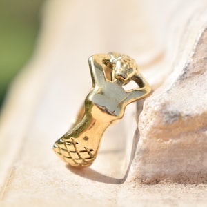 May include: A gold-colored ring in the shape of a mermaid. The mermaid is depicted with her arms behind her head, and the tail has a crosshatch pattern. The ring is set against a neutral background.
