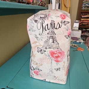 May include: A white fabric sewing machine cover with a pink and black Eiffel Tower design. The cover has the word "Paris" written in black cursive lettering.