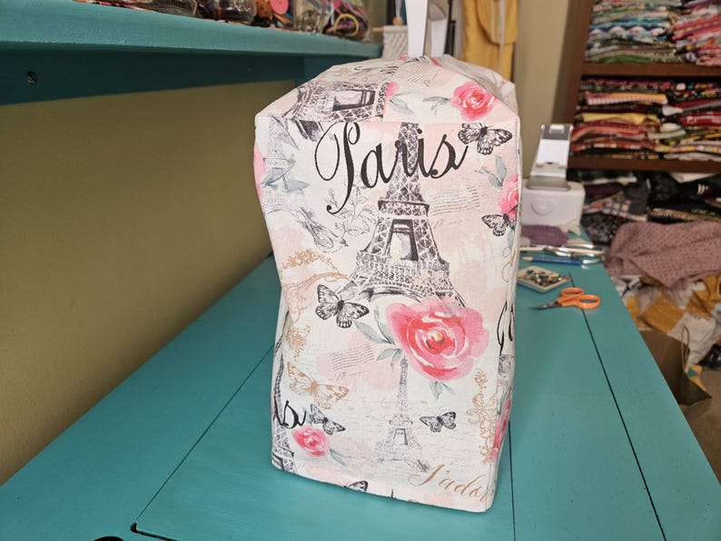 May include: A white fabric sewing machine cover with a pink and black Eiffel Tower design. The cover has the word "Paris" written in black cursive lettering.