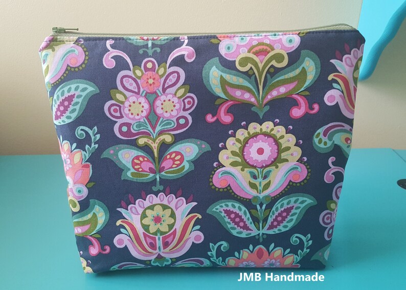 Pode incluir: Uma bolsa azul escura com z&iacute;per com um padr&atilde;o floral em tons de rosa, verde e amarelo. A bolsa tem um z&iacute;per e o texto "JMB Handmade" est&aacute; impresso na parte inferior.