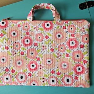 K&ouml;nnte beinhalten: Gesteppte Stofftasche mit floralem Muster. Die Tasche hat einen wei&szlig;en Hintergrund mit gro&szlig;en rosa und pfirsichfarbenen Blumen, kleinen roten Blumen und gr&uuml;nen Bl&auml;ttern. Sie hat einen passenden Griff und einen Rei&szlig;verschluss.