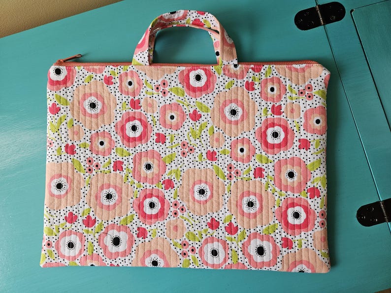 K&ouml;nnte beinhalten: Gesteppte Stofftasche mit floralem Muster. Die Tasche hat einen wei&szlig;en Hintergrund mit gro&szlig;en rosa und pfirsichfarbenen Blumen, kleinen roten Blumen und gr&uuml;nen Bl&auml;ttern. Sie hat einen passenden Griff und einen Rei&szlig;verschluss.