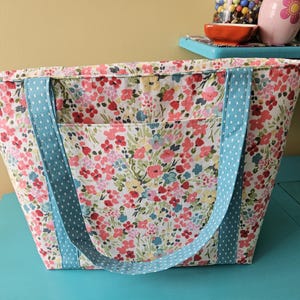 May include: A floral print tote bag with a light blue polka dot handle. The bag features a colorful pattern of pink, red, and yellow flowers on a white background. The tote has a front pocket and is sitting on a turquoise surface.