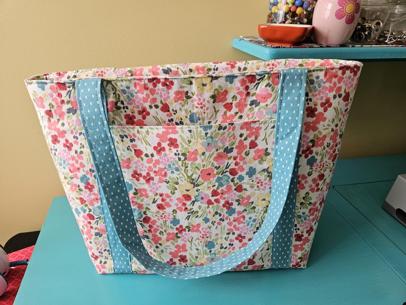 May include: A floral print tote bag with a light blue polka dot handle. The bag features a colorful pattern of pink, red, and yellow flowers on a white background. The tote has a front pocket and is sitting on a turquoise surface.