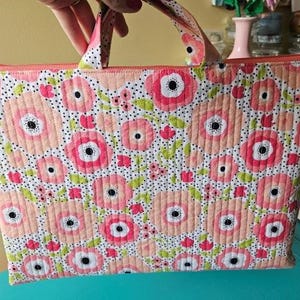 K&ouml;nnte beinhalten: Eine gesteppte Tragetasche mit floralem Muster. Die Tasche zeigt ein sich wiederholendes Design aus rosa und pfirsichfarbenen Blumen mit schwarzen Zentren und gr&uuml;nen Bl&auml;ttern auf wei&szlig;em Grund. Sie hat einen Rei&szlig;verschluss und passende Griffe.