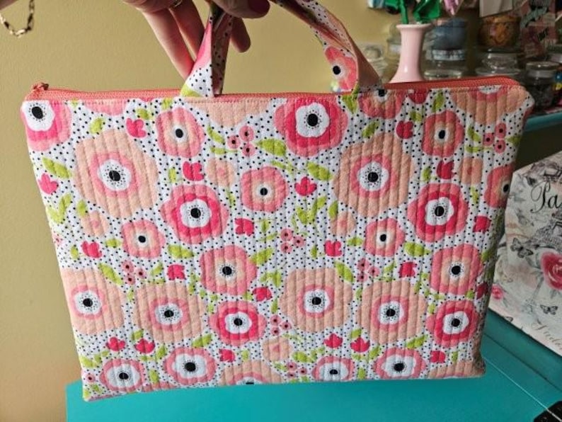 K&ouml;nnte beinhalten: Eine gesteppte Tragetasche mit floralem Muster. Die Tasche zeigt ein sich wiederholendes Design aus rosa und pfirsichfarbenen Blumen mit schwarzen Zentren und gr&uuml;nen Bl&auml;ttern auf wei&szlig;em Grund. Sie hat einen Rei&szlig;verschluss und passende Griffe.