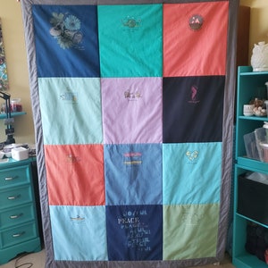 May include: A handmade quilt composed of colorful squares, each featuring a different graphic design and text. The quilt has a gray border and is held up by unseen hands. The squares are various colors, including blue, green, pink, and coral.