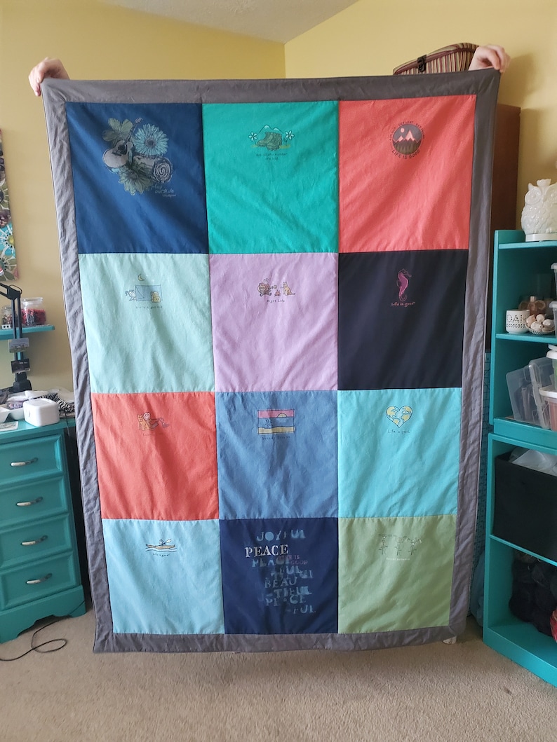 May include: A handmade quilt composed of colorful squares, each featuring a different graphic design and text. The quilt has a gray border and is held up by unseen hands. The squares are various colors, including blue, green, pink, and coral.