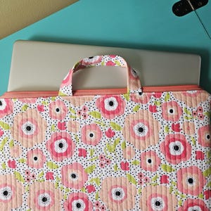 K&ouml;nnte beinhalten: Eine Laptop-H&uuml;lle mit Blumenmuster und Griff. Die H&uuml;lle hat ein gestepptes Design mit gro&szlig;en rosa und pfirsichfarbenen Blumen, schwarzen Tupfen und gr&uuml;nen Bl&auml;ttern auf wei&szlig;em Hintergrund. Sie hat einen Pfirsichfarbenen Rei&szlig;verschluss und Griff.