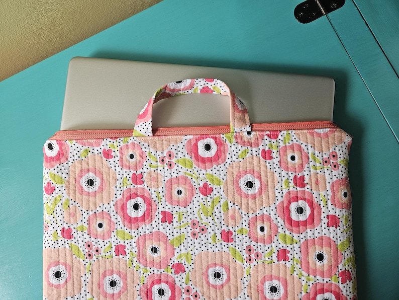 K&ouml;nnte beinhalten: Eine Laptop-H&uuml;lle mit Blumenmuster und Griff. Die H&uuml;lle hat ein gestepptes Design mit gro&szlig;en rosa und pfirsichfarbenen Blumen, schwarzen Tupfen und gr&uuml;nen Bl&auml;ttern auf wei&szlig;em Hintergrund. Sie hat einen Pfirsichfarbenen Rei&szlig;verschluss und Griff.