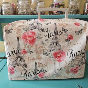 May include: A white sewing machine cover with a Paris theme. The cover features a repeating pattern of the Eiffel Tower, pink roses, butterflies, and the words "Paris" and "J'adore".
