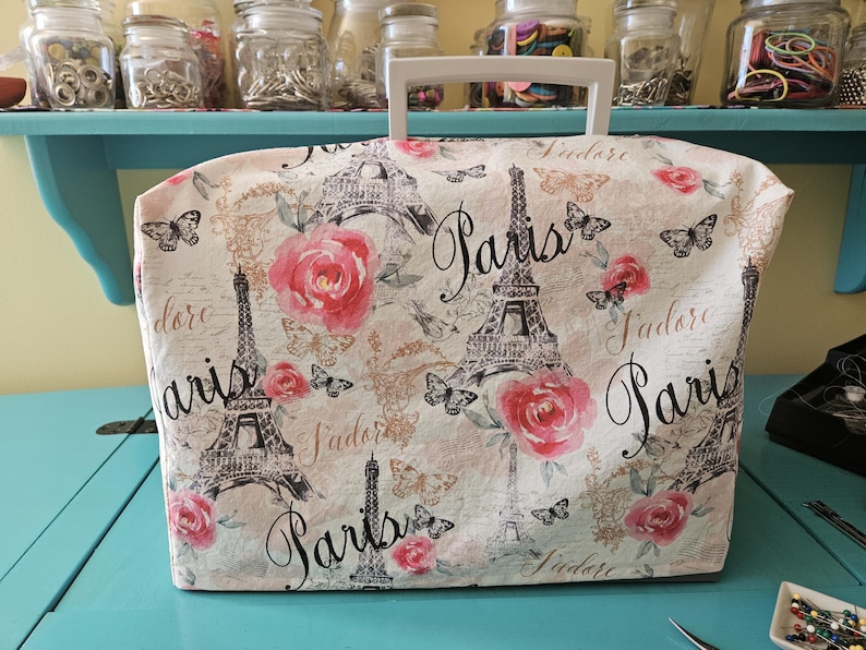 May include: A white sewing machine cover with a Paris theme. The cover features a repeating pattern of the Eiffel Tower, pink roses, butterflies, and the words "Paris" and "J'adore".