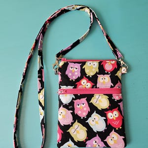 Peut inclure: Un sac bandouli&egrave;re noir avec une fermeture &eacute;clair et une sangle roses. Le sac est orn&eacute; de motifs de hiboux color&eacute;s. La sangle et la fermeture &eacute;clair sont roses. Le fond est de couleur turquoise unie.