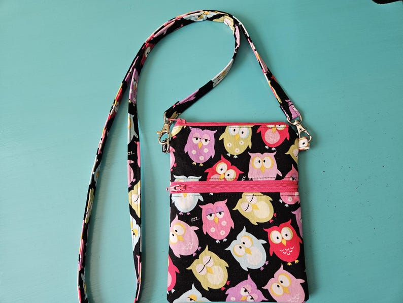 Peut inclure: Un sac bandouli&egrave;re noir avec une fermeture &eacute;clair et une sangle roses. Le sac est orn&eacute; de motifs de hiboux color&eacute;s. La sangle et la fermeture &eacute;clair sont roses. Le fond est de couleur turquoise unie.