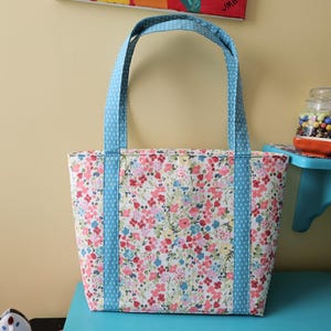 May include: A floral tote bag with light blue handles and accents. The bag has a pattern of pink, red, and blue flowers on a white background. The handles and side accents are light blue with white polka dots.