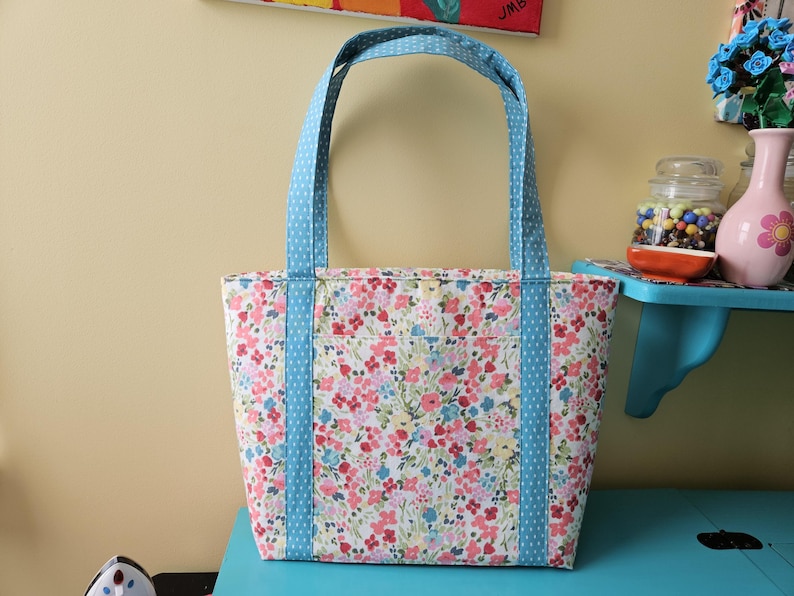 May include: A floral tote bag with light blue handles and accents. The bag has a pattern of pink, red, and blue flowers on a white background. The handles and side accents are light blue with white polka dots.