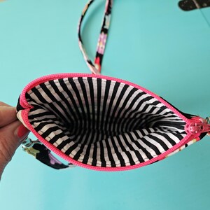 Peut inclure: Petite pochette &agrave; imprim&eacute; floral noir, fermeture &eacute;clair rose et int&eacute;rieur &agrave; rayures noires et blanches. La pochette est ouverte, r&eacute;v&eacute;lant l'int&eacute;rieur. Elle a une bordure rose et une sangle assortie.
