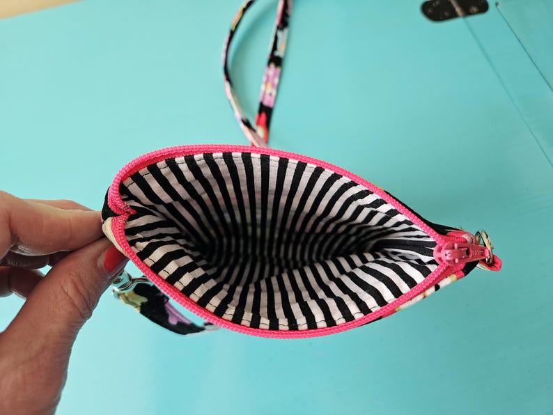 Peut inclure: Petite pochette &agrave; imprim&eacute; floral noir, fermeture &eacute;clair rose et int&eacute;rieur &agrave; rayures noires et blanches. La pochette est ouverte, r&eacute;v&eacute;lant l'int&eacute;rieur. Elle a une bordure rose et une sangle assortie.